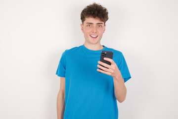 young caucasian handsome man with curly hair wearing blue T-shirt against white studio background holds mobile phone in hands and rejoices positive news, uses modern cellular