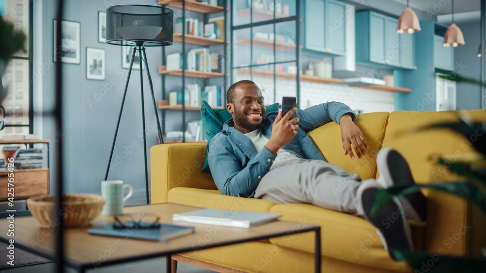 Leinwandbilder excited black african american man using smartphone while resting on a sofa in living room. happy ma
