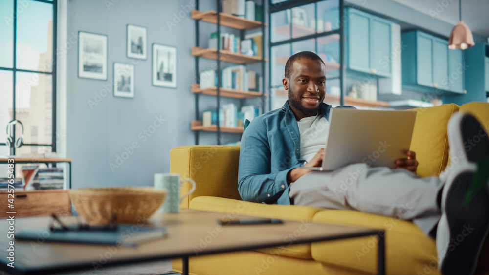 Poster handsome black african american man working on laptop computer while lying on a sofa in cozy living 
