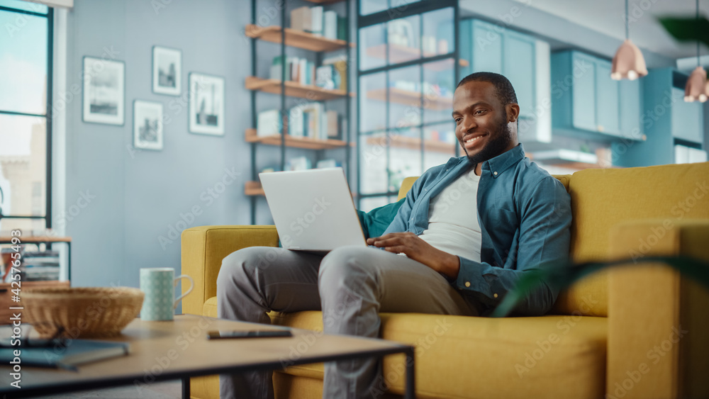 Poster happy black african american man working on laptop computer while sitting on a sofa in cozy living r