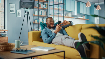 Excited Black African American Man Using Smartphone while Resting on a Sofa in Living Room. Happy Man Smiling at Home and Chatting to Colleagues and Clients Over the Internet. Using Social Networks. - obrazy, fototapety, plakaty