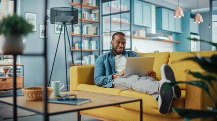 Handsome Black African American Man Working on Laptop Computer while Lying on a Sofa in Cozy Living Room. Freelancer Working From Home. Browsing Internet, Using Social Networks, Having Fun in Flat.