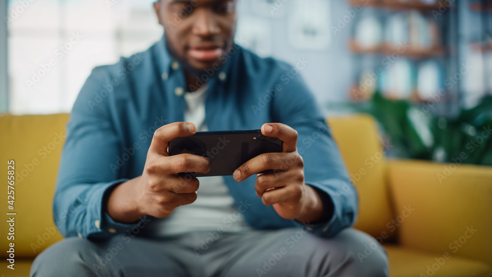 Poster happy black african american man is playing video game on smartphone app while sitting on a sofa in 