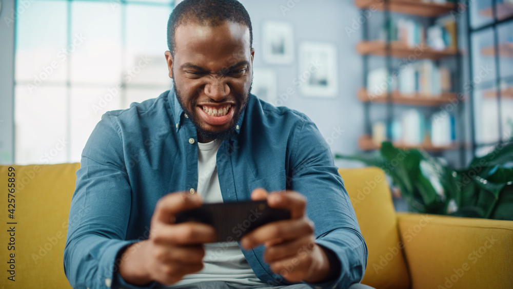Poster angry black african american man playing video game on smartphone app while sitting on a sofa in liv