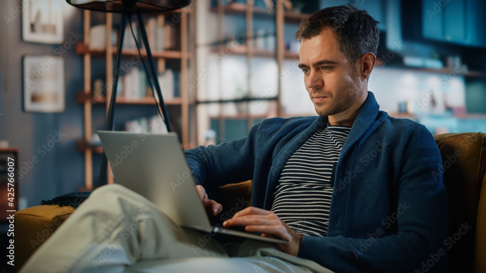 Aufkleber Handsome Caucasian Man Working on Laptop Computer while Sitting on a Sofa in Stylish Cozy Living Room. Freelancer Working From Home. Browsing Internet, Using Social Networks, Having Fun in Flat.