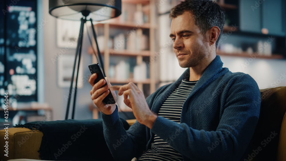 Aufkleber Excited Caucasian Man Using Smartphone while Resting on a Sofa in Stylish Living Room. Happy Man Smiling at Home and Chatting to Colleagues and Clients Over the Internet. Using Social Networks.