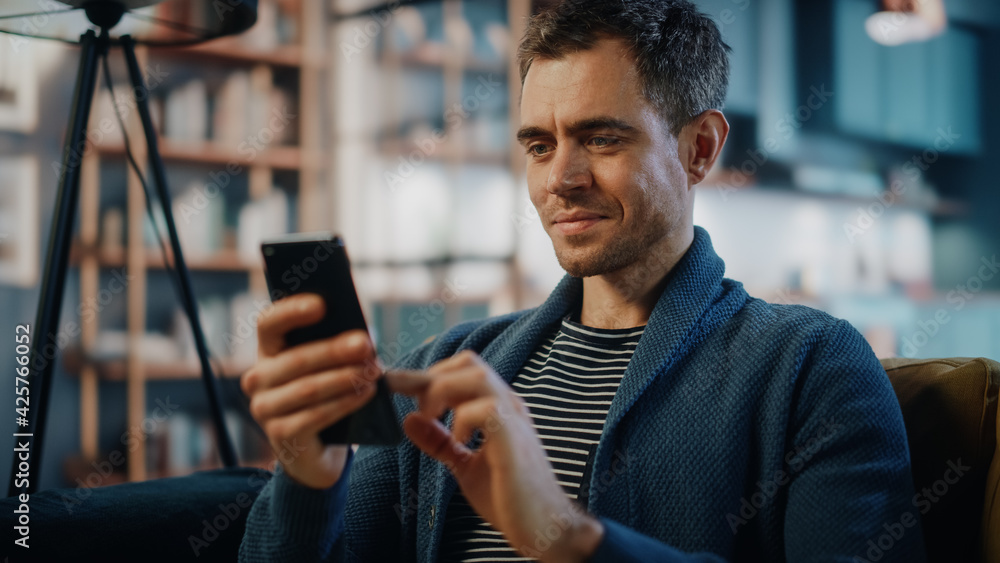 Aufkleber Excited Caucasian Man Using Smartphone while Resting on a Sofa in Stylish Living Room. Happy Man Smiling at Home and Chatting to Colleagues and Clients Over the Internet. Using Social Networks.