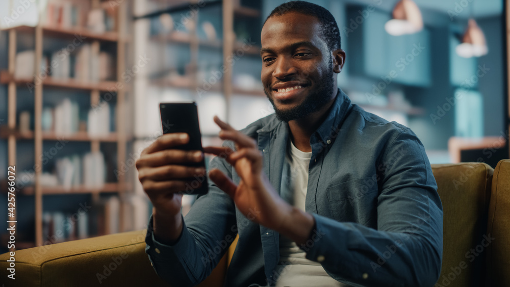 Aufkleber Excited Black African American Man Using Smartphone while Resting on a Sofa in Living Room. Happy Man Smiling at Home and Chatting to Colleagues and Clients Over the Internet. Using Social Networks.