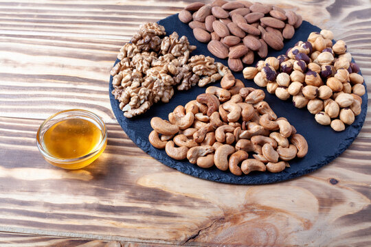 Various nuts sorted on round stone plate with honey glass bowl and honet dipper. Mixed nuts on wooden table. Black stone plate on wooden background.