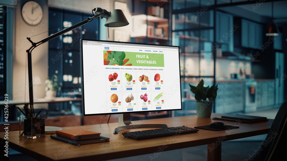 Poster Shot of a Desktop Computer with Food App User Interface Developing Software Standing on the Wooden Desk in the Creative Cozy Living Room. In the Background Warm Evening Lighting and Open Space Studio.