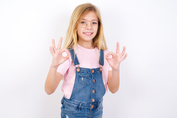 beautiful caucasian little girl wearing denim jeans overall over white background showing both hands with fingers in OK sign. Approval or recommending concept