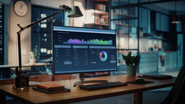 Shot of a Desktop Computer with Financial Market Analysis Application Standing on the Wooden Desk in the Creative Cozy Living Room. In the Background Warm Evening Lighting and Open Space Studio.