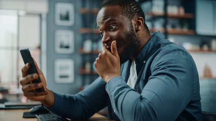 Shocked Black African American Man Using Smartphone while Sitting at Table in Living Room. Man...