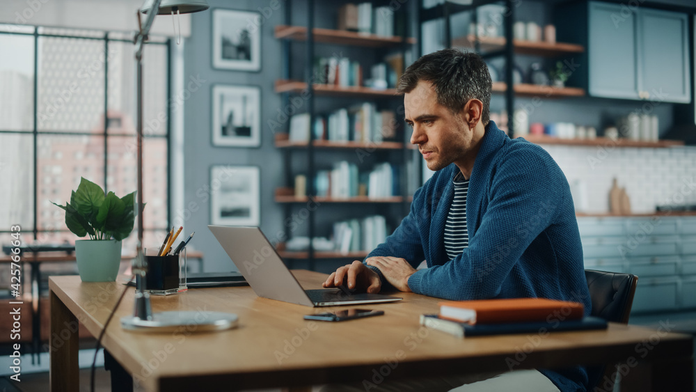 Poster Handsome Caucasian Man Working on Laptop Computer while Sitting Behind Desk in Cozy Living Room. Freelancer Working From Home. Browsing Internet, Using Social Networks, Having Fun in Flat.