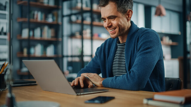 Handsome Caucasian Man Working On Laptop Computer While Sitting Behind Desk In Cozy Living Room. Freelancer Working From Home. Browsing Internet, Using Social Networks, Having Fun In Flat.