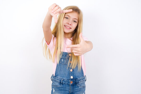 beautiful caucasian little girl wearing denim jeans overall over white background making finger frame with hands. Creativity and photography concept.