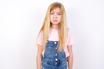 Displeased upset beautiful caucasian little girl wearing denim jeans overall over white background frowns face as going to cry, being discontent and unhappy as can't achieve goals,  Disappointed girl