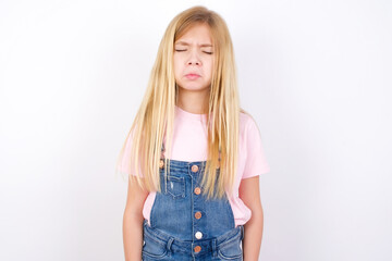 Fototapeta premium Dismal gloomy rejected beautiful caucasian little girl wearing denim jeans overall over white background has problems and difficulties, curves lower lip and closes eyes in despair, being in depression