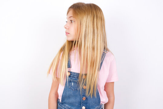Close up side profile photo beautiful caucasian little girl wearing denim jeans overall over white background not smiling attentive listen concentrated