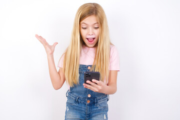 beautiful caucasian little girl wearing denim jeans overall over white background holding in hands cell reading browsing news