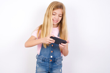 beautiful caucasian little girl wearing denim jeans overall over white background holding in hands cell playing video games or chatting