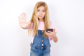 beautiful caucasian little girl wearing denim jeans overall over white wall using and texting with smartphone with open hand doing stop sign with serious and confident expression, defense gesture