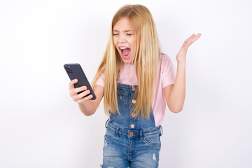Fototapeta premium Angry beautiful caucasian little girl wearing denim jeans overall over white background screaming on the phone, having an argument with an employee. Troubles at work.