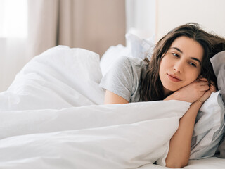 Obraz na płótnie Canvas Cozy morning. Sunday relaxation. Home rest. Peaceful thoughtful daydreaming brunette woman enjoying lying in bed alone with white blanket in light bedroom.