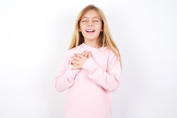 Beautiful caucasian blonde little girl wearing pink sweater expresses happines, laughs pleasantly, keeps hands on heart