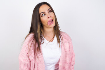 Fototapeta premium Young beautiful caucasian woman wearing pink knitted jacket over white wall showing grimace face crossing eyes and showing tongue. Being funny and crazy