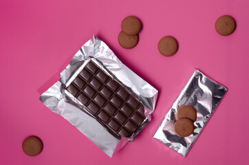 One opened dark chocolate bar on the silver foil, sweet cookies and whole chocolate bar on the bright pink table