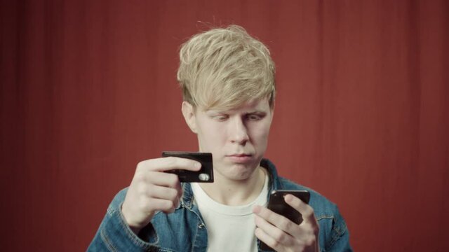 Portrait of young british man holding phone entering pincode with credit card, doing shopping online. Student using plastic card making transaction using mobile banking application. 