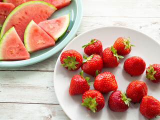 Fresh strawberries on a white plate and juicy watermelon slices on a green plate, white wooden background, closeup

