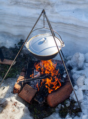 Over the fire hangs a pot in which to cook food. On a hook on a tripod, steam comes out of the pan....