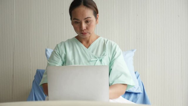 Medium shot portrait of middle-gaed adult Asian woman wearing patient outfit, sitting on bed, using laptop computer, working in hospital ward. Busy life of employee duting sick leave concept.