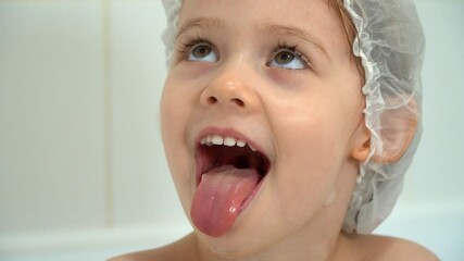 Close up of little funny girl bathes in bath, looks at camera, smiles and laughs. Happy child bathes with soap bubbles in white bathroom. Baby bathing - Powered by Adobe