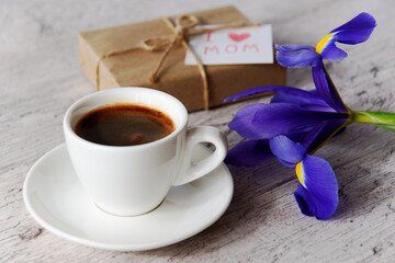 White cup of coffee, gift box with message for mom and iris flower on gray wood table. Mother's Day holiday concept.
