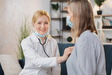 Professional female doctor or nurse doing the auscultation of her senior lady patient at home, listening the heart beat and smiling to camera. Doctor taking the heartbeat of patient.