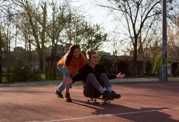 Two young female friends hang out in the city streets. One girl is sitting on the skateboard while the other one is pushing her around.