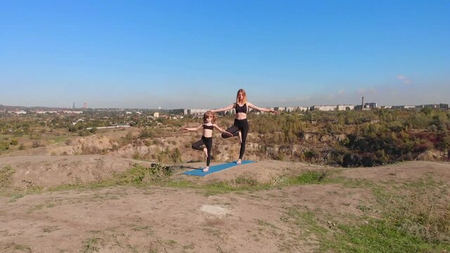 Aerial drone view of slender young mother do yoga exercises with child daughter on high hill by lake in morning, far from bustle of city, set good example and instill in child healthy habits