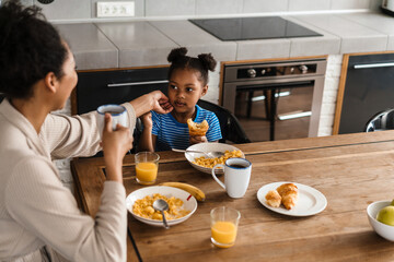 Happy black mother and daughter having breakfast at home kitchen