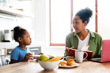 Black smiling woman working while having breakfast with her daughter