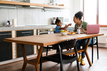 Black smiling woman working while having breakfast with her daughter