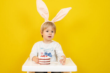 Beautiful baby boy wearing rabbit headband sitting and holding a cake isolated over yellow background.