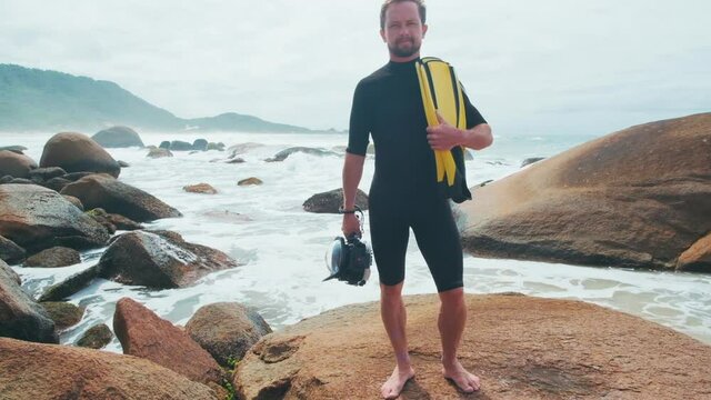 Surf photographer. Portrait of caucasian surfing photographer standing on the rocky coast with fins and underwater housing in hands