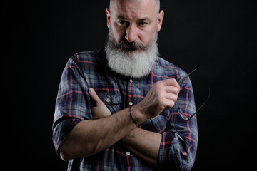 Studio portrait of mature bearded man in casual clothes with eyeglasses in hand looking directly at camera, informal lifestyle, black backdrop.