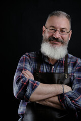 Portrait of handsome mature bearded man dressed leather apron isolated on black background, caucasian workman with beard smiling