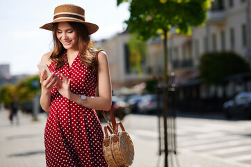 Beautiful young woman using smartphone and walks in the summer city wearing straw hat and beautiful...