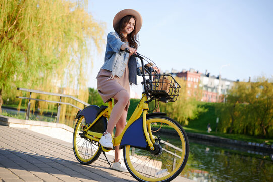 Beautiful young smiling woman in a summer outfit strolls around the city and rides rental bicycle by the river