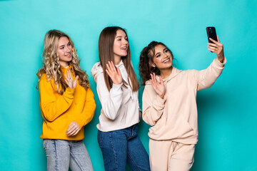 Young three multiethnic women friends make video call on phone posing isolated over turquoise background